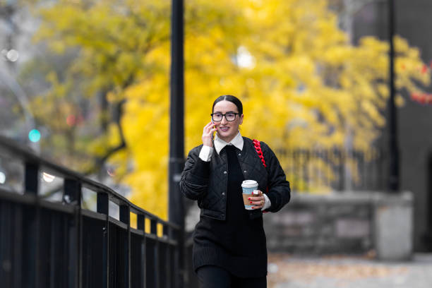 A stylish woman in black glasses, holding a coffee and phone, walks outdoors in a fall setting.